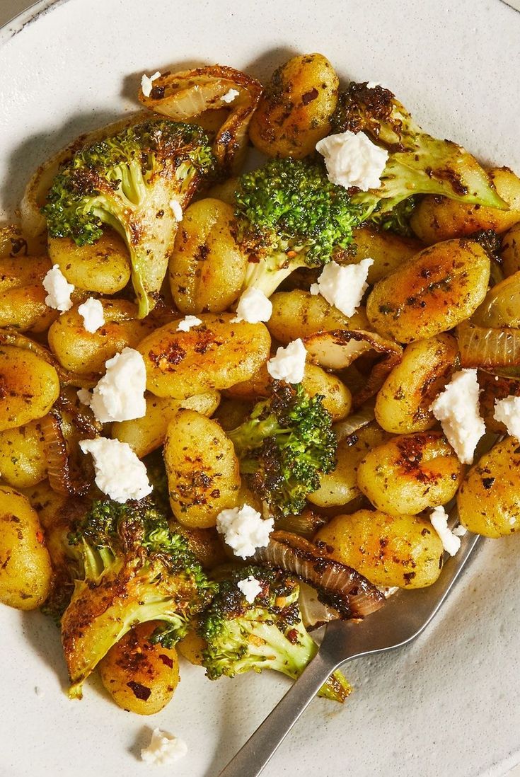 a white plate topped with potatoes and broccoli covered in feta next to a fork