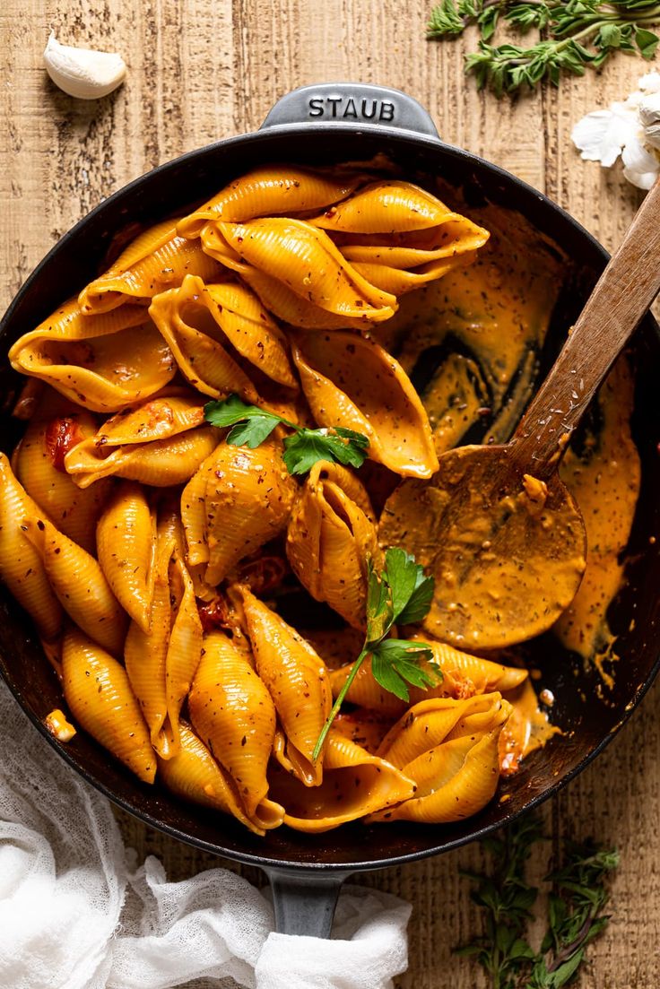 a skillet filled with pasta and sauce on top of a wooden table next to garlic