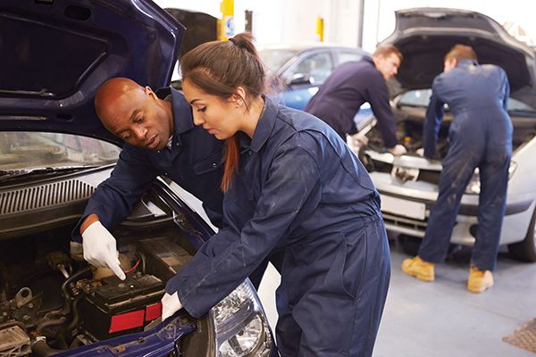 two people working on an automobile in a garage with the hood open and one man looking at the engine