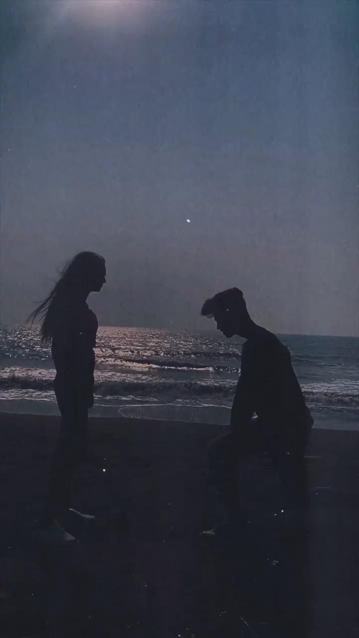 two people are standing on the beach at night, one is looking at the water