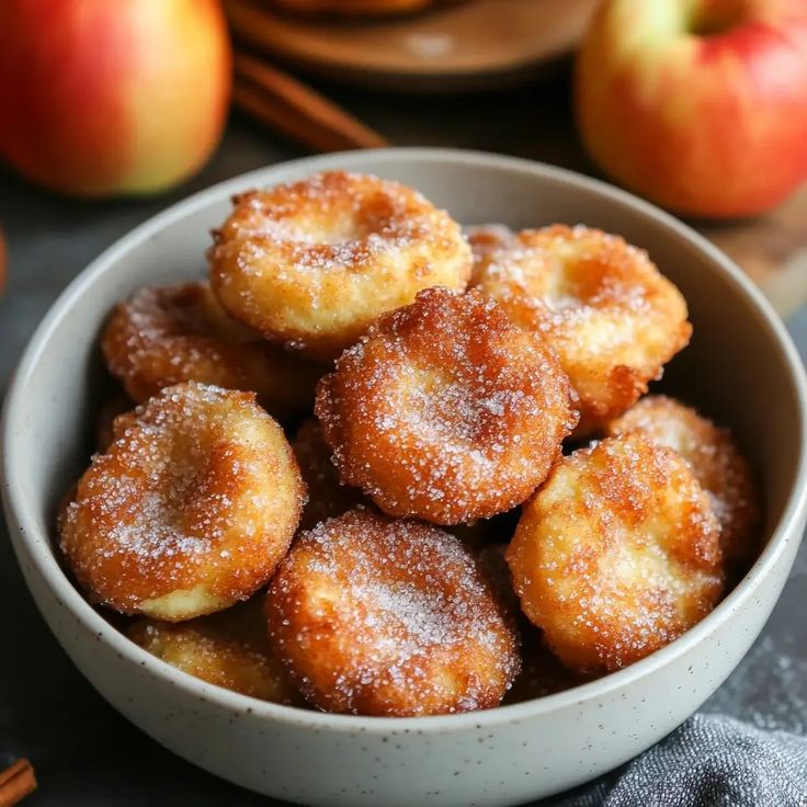 a bowl filled with sugar covered donuts on top of a table next to apples