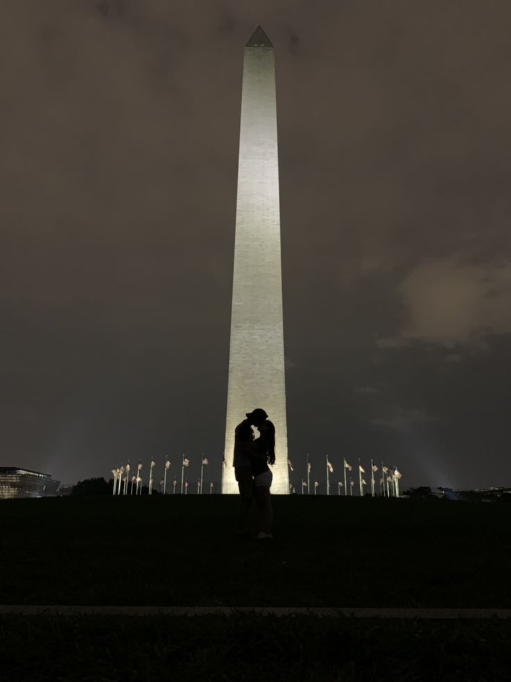 two people standing in front of the washington monument at night with their arms around each other