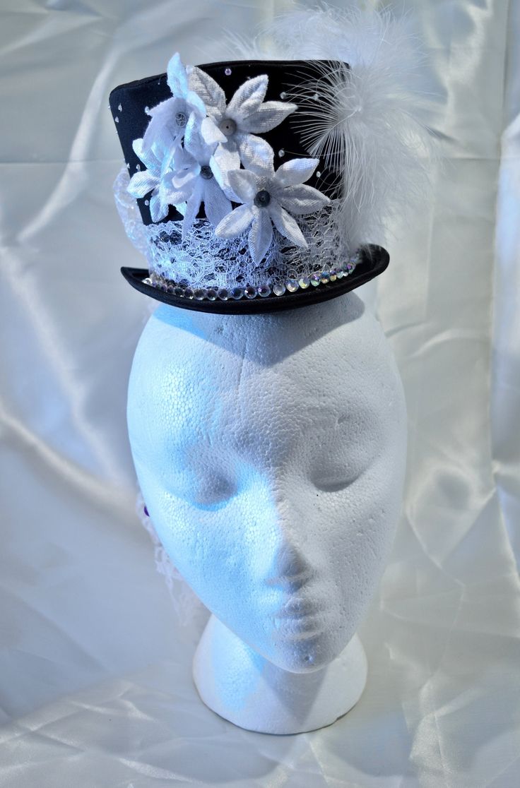 This hat is excellent for making a statement, its neautral colors allow you to build it into a focal point with any outfit. This satin miniature top hat can be worn with a headband or affixed to the wearers head via small loops inside the hat. Delicate white lace makes up the band, creating a stark contrast with the satin hat and forming a generous bow and waterfall tail in the back. The front of the band is decorated with a line of large rhinestones. Smaller rhinestones scatter thier glittery light around the rest of the hat. The front of the hat features small white velvet poinsettia flowers, with small gear and cog centers. The side features white feathers with a black and white cameo. Whimsical Fitted Top Hat With Short Brim, Handmade Fitted High Crown Mini Hats, Handmade Fitted Mini Hat With High Crown, Whimsical Adjustable Top Hat For Costume, Brimmed Party Costume Hats And Headpieces, Whimsical Party Hats, Handmade White Mini Hats For Party, Fitted Tall Crown Hat For Party, Elegant Adjustable Fascinator For Festivals