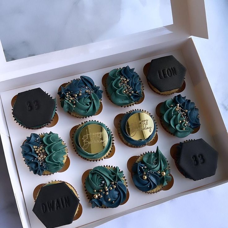 twelve decorated cupcakes in a white box with black and gold decorations on them