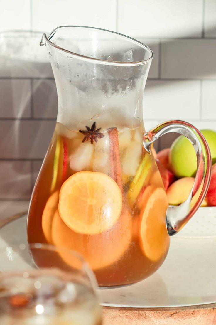 a pitcher filled with liquid sitting on top of a table next to apples and oranges