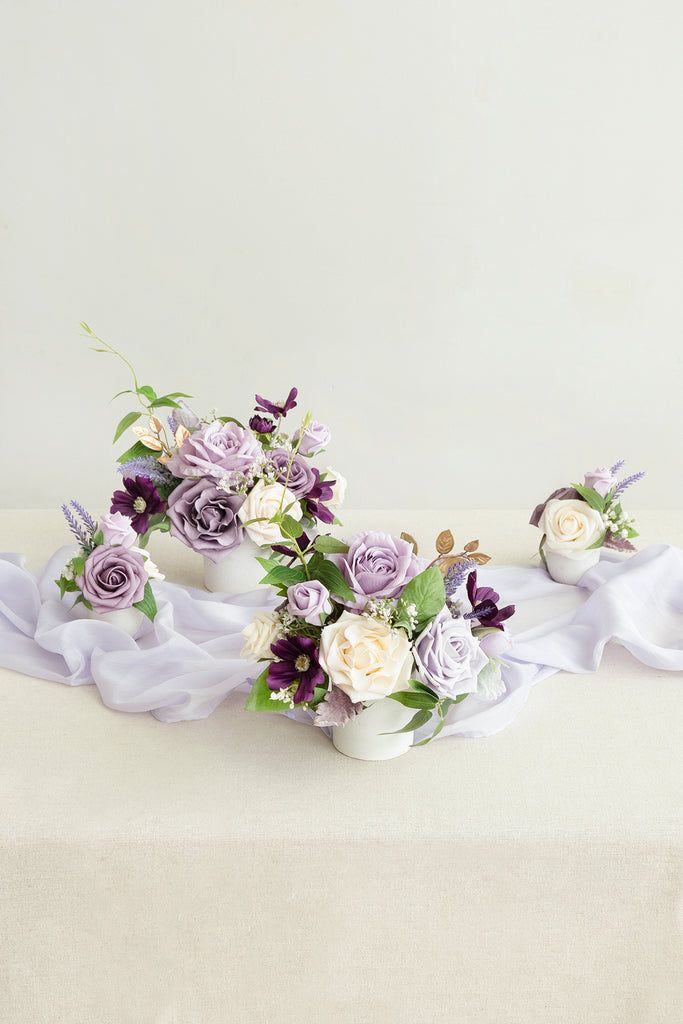 two white vases with purple flowers and greenery on a tablecloth draped around them