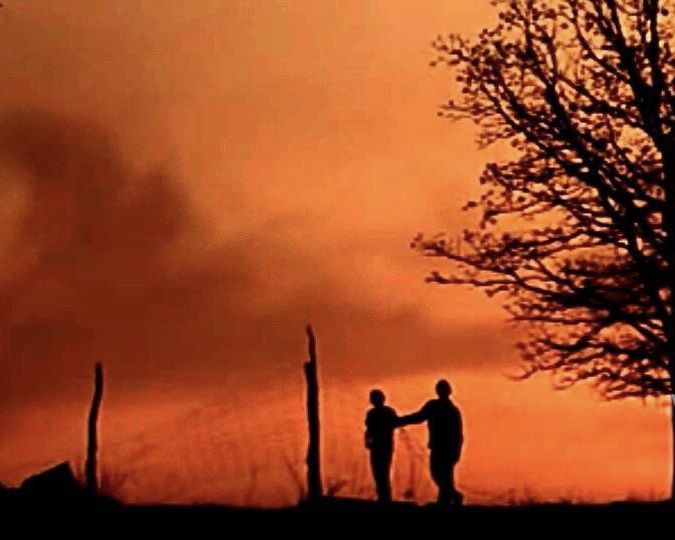 two people standing next to each other in front of an orange sky with the sun setting behind them