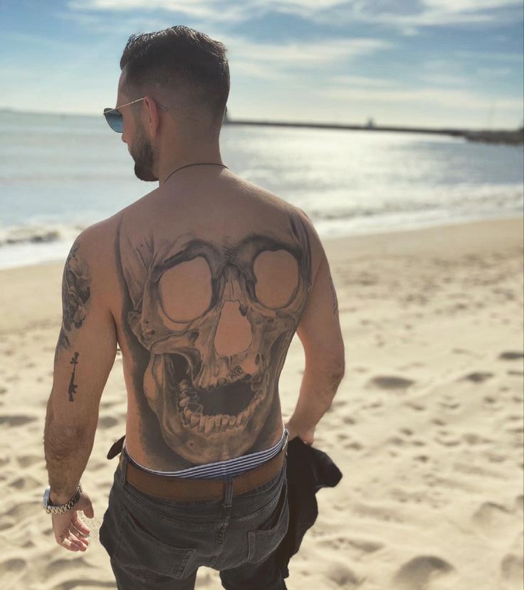 a man standing on top of a beach next to the ocean with a skull tattooed on his back