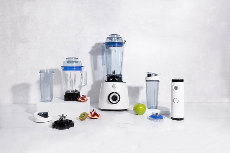 several blenders are lined up on a white surface with fruits and vegetables around them