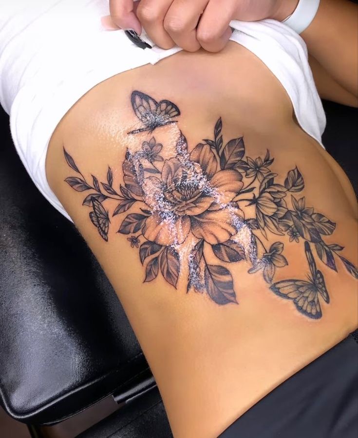 a woman's stomach with flowers and butterflies on the side, while she is getting her tattoo done