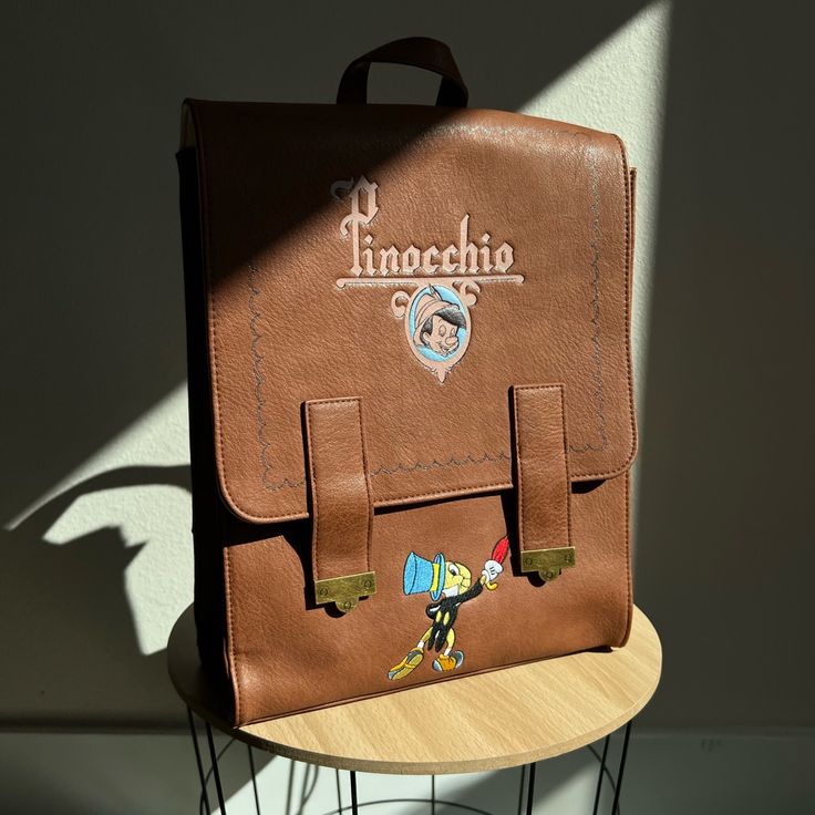 Loungefly Disney's Pinocchio Mini Satchel Backpack Is A Boxlunch Exclusive & Is Brand New But No Tags. Features Include Main Compartment, Interior Pocket, Bronze Hardware, Embroidered Jimmy Cricket, Debased Characters On The Sides, A Logo Graphic On The Front & Under The Flap Of Pinnochio's Hometown With The Stars Shining Brightly Above, Magnetic Closure Flap, Adjustable Straps. Measures 10.5 X 13.25 X 4.5" Themed Backpack With Case Included, Disney Backpack With Case For School, Disney Style Backpack Bag As Gift, Disney Style Backpack Bag For Gifts, Disney Backpack For School With Case Included, Disney Leather School Bags, Disney Rectangular Backpack For Everyday Use, Disney Themed Rectangular Backpack For Everyday Use, Disney Leather School Backpack