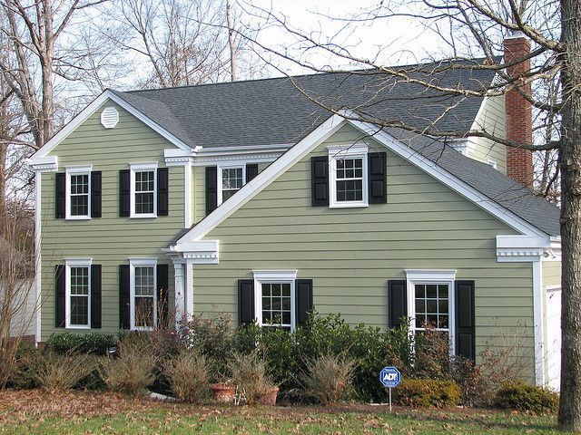 a green house with white trim and black shutters on the front, surrounded by trees