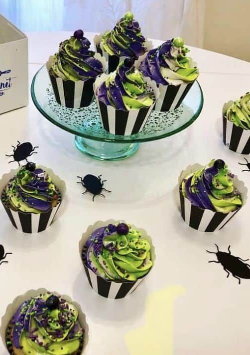 cupcakes decorated with green and purple frosting on a table next to a box of chocolate bugs