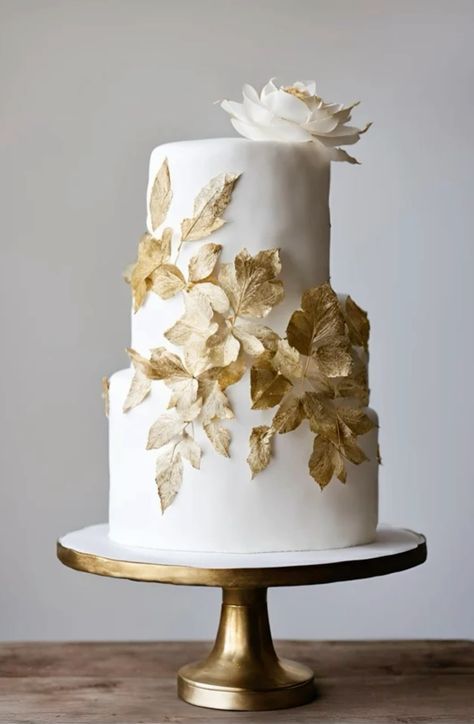 a white and gold wedding cake with leaves on top is sitting on a wooden table