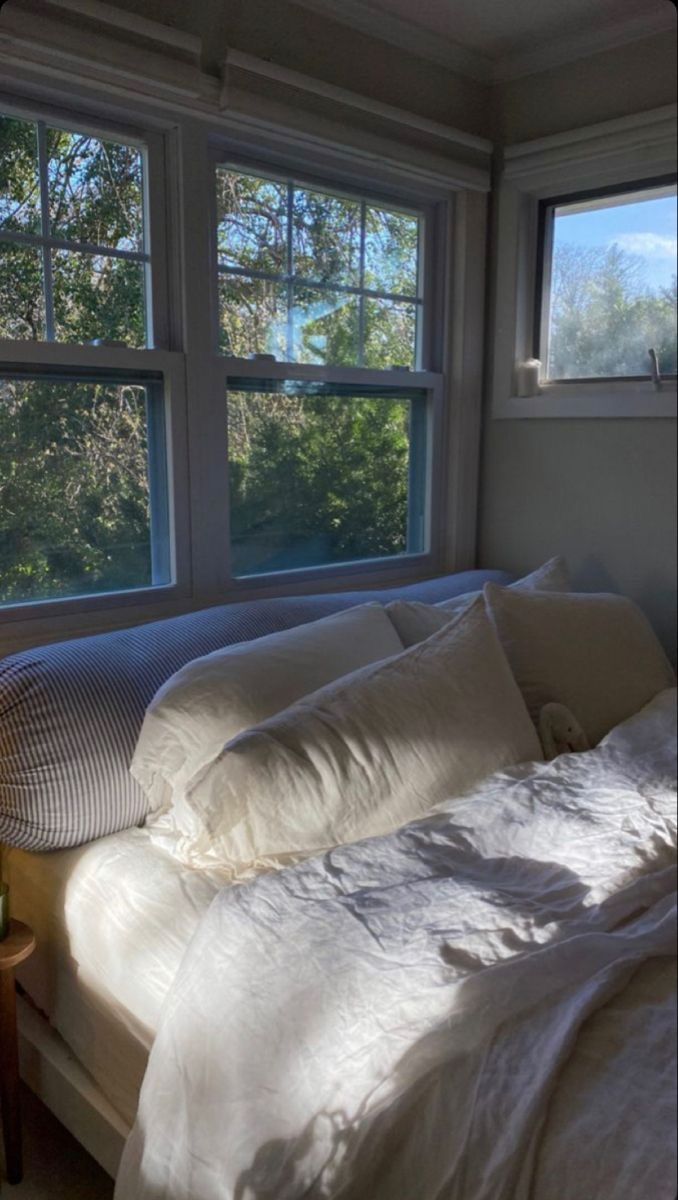 a bed with white sheets and pillows in front of two large windows that look out onto the trees