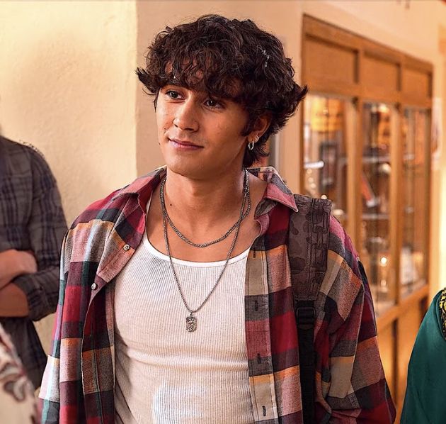 a young man with curly hair wearing a plaid shirt and carrying a backpack in his hand