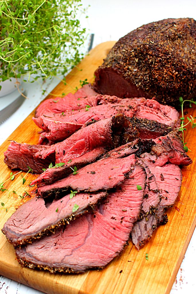 sliced roast beef sitting on a cutting board with herbs and seasoning next to it