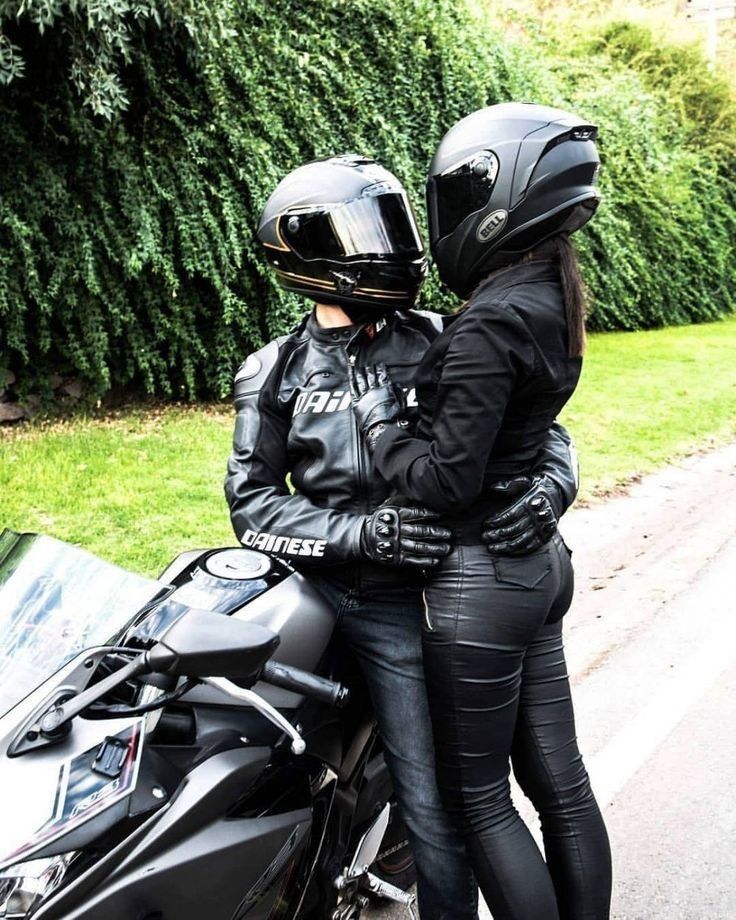 two people on a motorcycle wearing helmets and black leather pants, standing next to each other