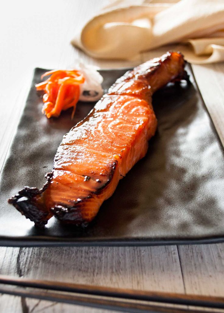 a piece of salmon on a black plate with carrots and chopsticks next to it