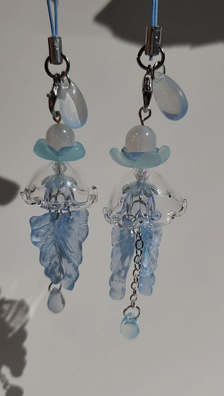 two blue and clear glass beads hanging from hooks on a white surface with shadow behind them