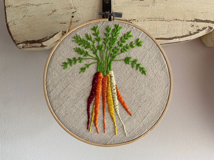 a cross stitch carrot with green leaves on it hanging from a wooden hoop next to a piece of wood