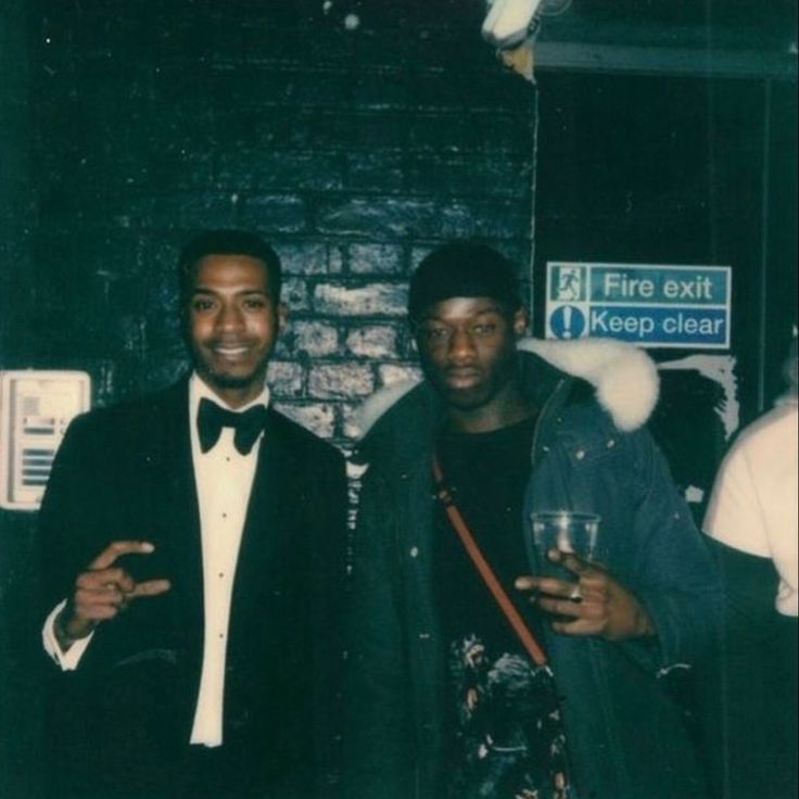 two men standing next to each other in front of a brick wall holding wine glasses