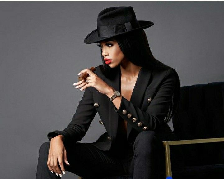 a woman in a black suit and hat sitting on a chair with her arms crossed