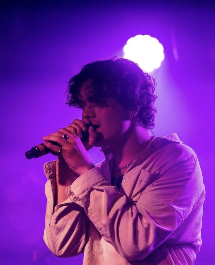 a young man holding a microphone in front of a purple stage light with his right hand up to his mouth