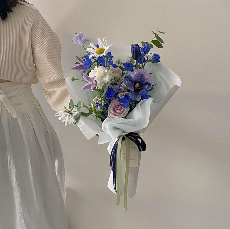 a woman holding a bouquet of blue and white flowers in her left hand while standing next to a wall