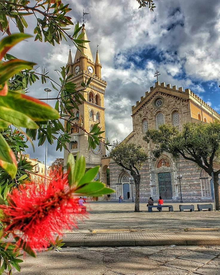 an old church with red flowers in the foreground and people sitting on benches near it