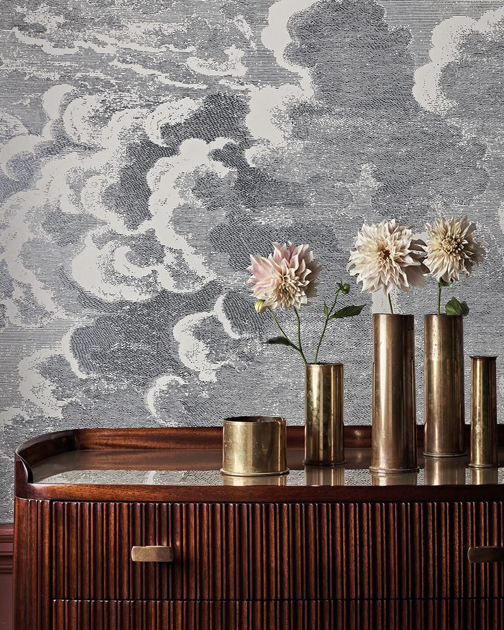 three brass vases with flowers in them on a wooden table next to a wall