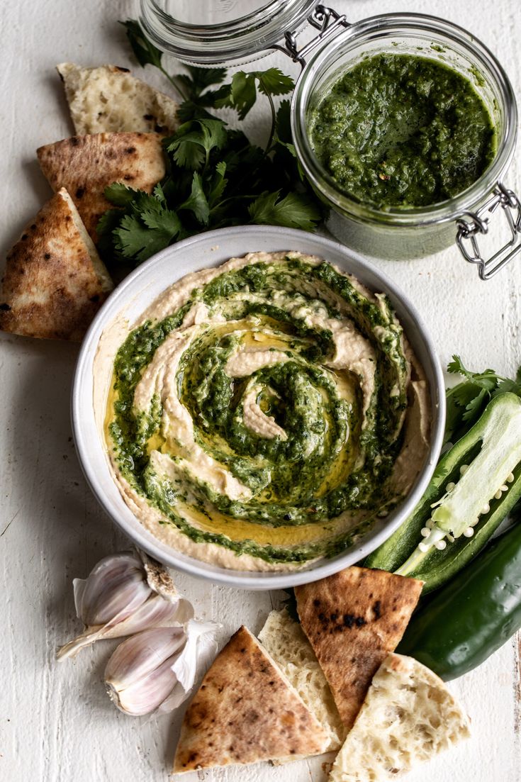 a white bowl filled with hummus next to some pita bread and green vegetables