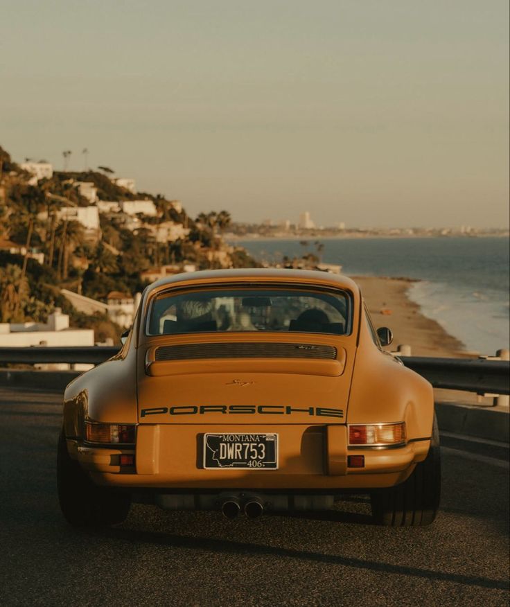 an orange porsche sits parked on the side of the road next to the ocean and houses