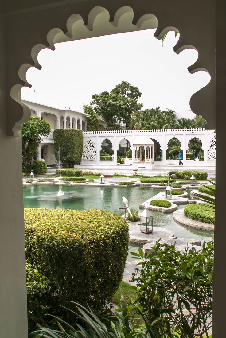 an outdoor garden with water and bushes in the foreground, surrounded by white arches