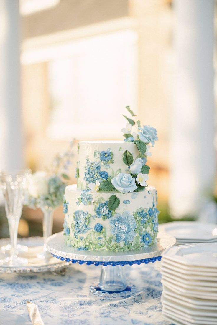 a wedding cake with blue flowers and greenery on the top is sitting on a table