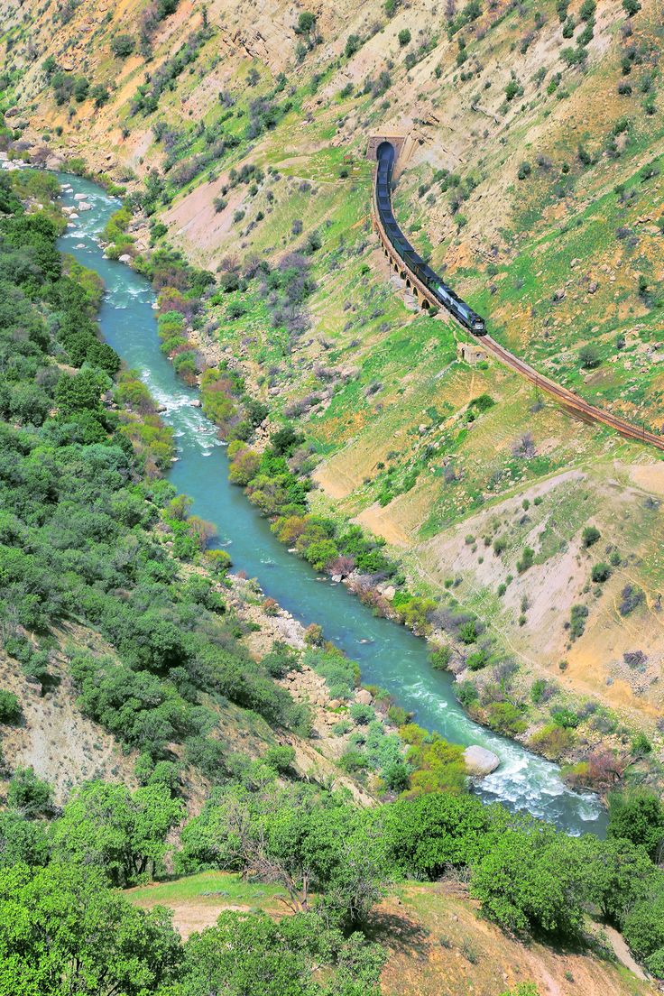 a train is going down the tracks near a river and trees in the mountainside