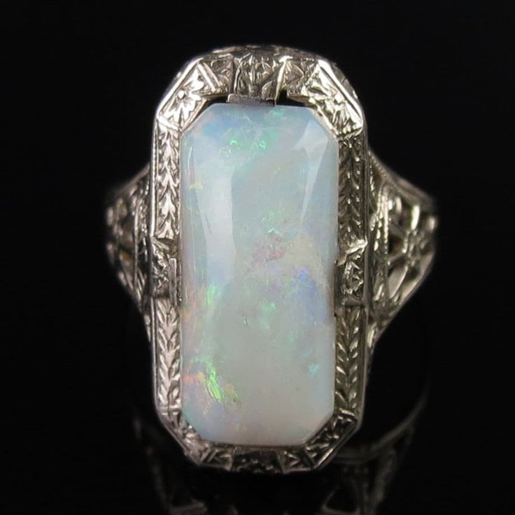 A natural white opal tops this ornate 14k white gold ring by famed jewelers, Ostby & Barton. A carved Art Deco design frames the 13mm x 6mm x 3.8mm cabochon. The white opal has a pinkish undertone with subtle flashes of greens and reds. A pierced filigree design with scrollwork and floral elements decorates the tapered shoulders. The inside of the ring is signed, "OB" (hallmark attributed to Ostby & Barton) followed by "14k" (white gold). The ring measures a petite size 4 (a pinky size for some) Classic Opal Ring Collectible, Elegant Hallmarked Opal Ring, Classic White Cabochon Opal Ring, Elegant Collectible Opal Ring, Classic Formal Opal Rings, Victorian Opal Ring For Formal Occasions, Heirloom Style Cabochon Opal Ring For Formal Occasions, Heirloom Cabochon Opal Ring For Formal Occasions, Heirloom Style Opal Cabochon Ring For Formal Occasions