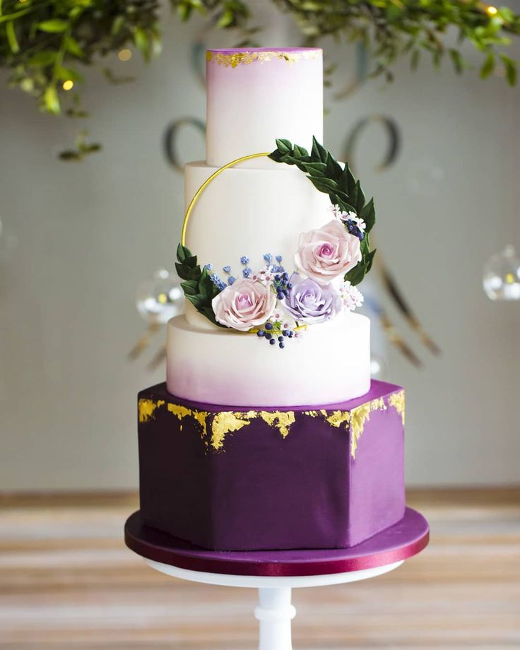 a three tiered cake with purple and white flowers on the top is decorated with greenery