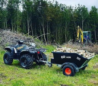 an atv pulling a trailer full of logs in the grass next to a pile of wood