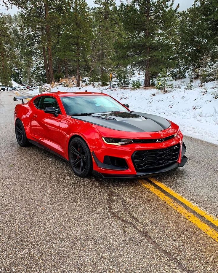 a red chevrolet camaro driving down a road in the winter with snow on the ground