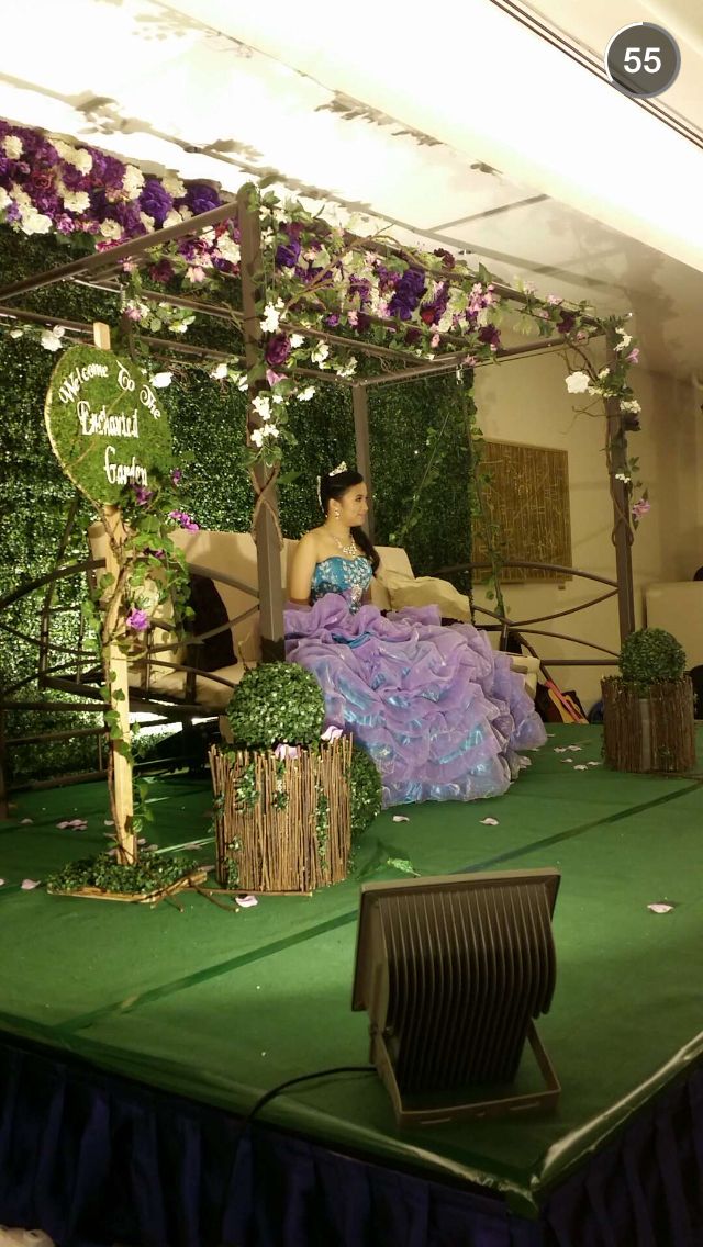 a woman in a purple dress sitting on a green stage with flowers and greenery