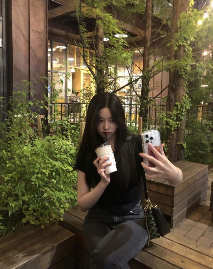 a woman sitting on a wooden bench taking a selfie with her cell phone while holding a cupcake