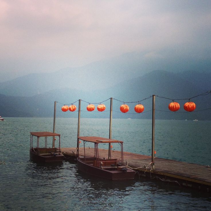 three boats tied to a dock with lanterns hanging from them over the water and mountains in the background