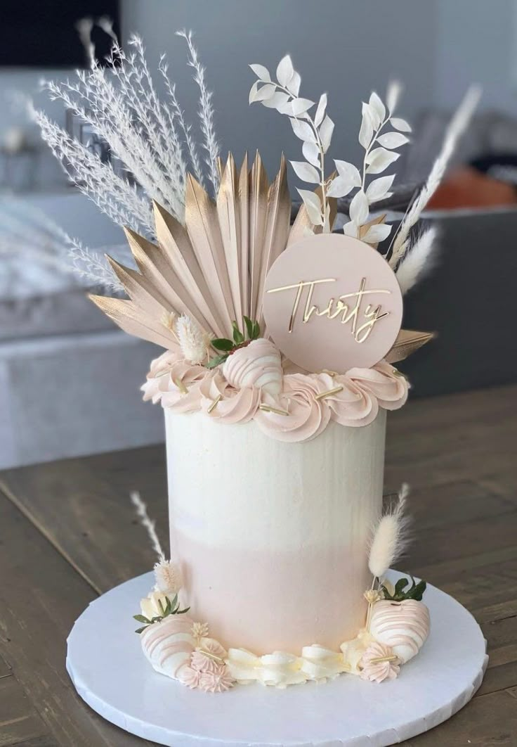 a white cake with pink flowers and feathers on top is sitting on a wooden table