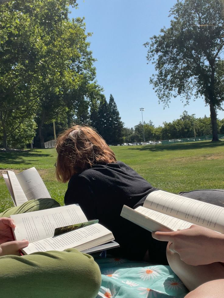 two people sitting on the grass with open books in their hands and one person laying down