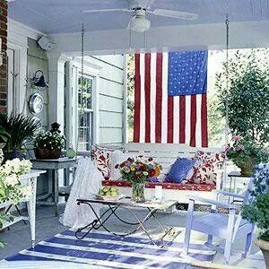 an american flag is hanging on the wall above a porch with white furniture and potted plants