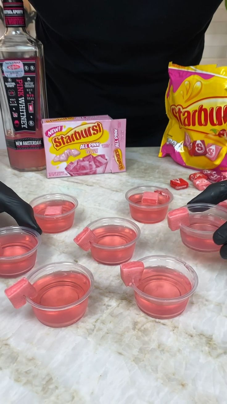 several pink cups are lined up on a table with black gloves and candy bar in the background