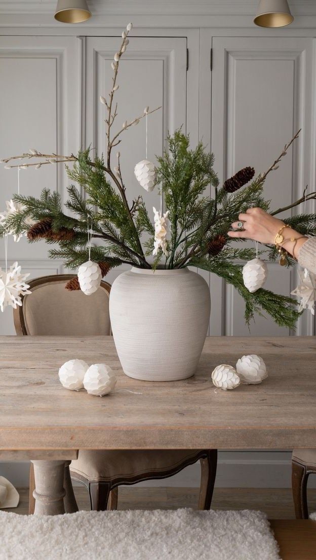 a woman arranging christmas decorations in a white vase on top of a wooden dining table