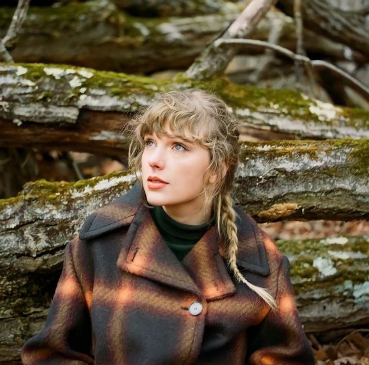 a woman with braids standing in front of some trees and mossy branches, wearing a coat