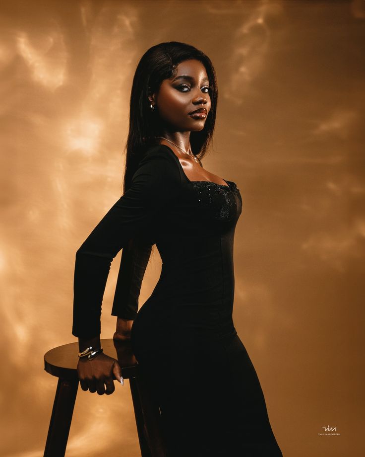 a woman in a black dress is posing for a photo with a stool and clouds behind her